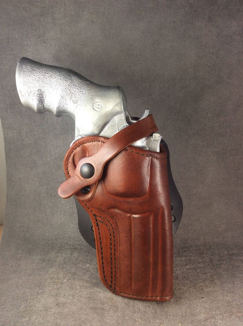 May include: A brown leather holster with a silver handgun inside. The holster has a strap with a snap closure and is designed for concealed carry.