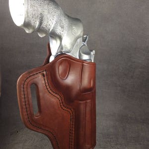 May include: A brown leather holster with a silver revolver inside. The holster has a belt loop and a stitched design.