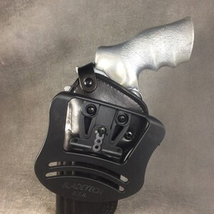 May include: Black leather holster with a black plastic frame and a silver gun. The holster has a Blade-Tech logo and says "U.S.A."