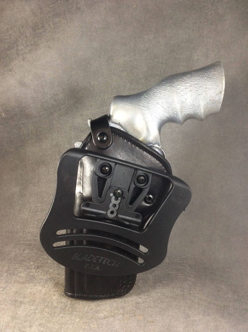 May include: Black leather holster with a black plastic frame and a silver gun. The holster has a Blade-Tech logo and says "U.S.A."