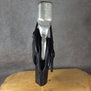 Op de afbeelding: Een zilveren metalen voorwerp wordt vastgehouden in een zwarte leren holster. De holster is met een messing pin aan een houten oppervlak bevestigd. De achtergrond is effen grijs.