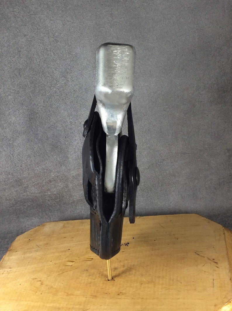 Op de afbeelding: Een zilveren metalen voorwerp wordt vastgehouden in een zwarte leren holster. De holster is met een messing pin aan een houten oppervlak bevestigd. De achtergrond is effen grijs.