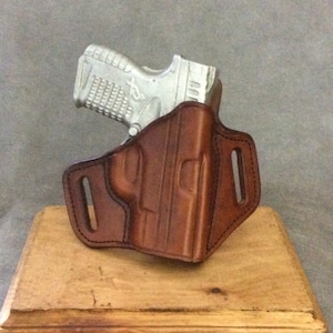 May include: A brown leather holster for a handgun. The holster has a belt loop and a snap closure. The holster is displayed on a wooden stand.