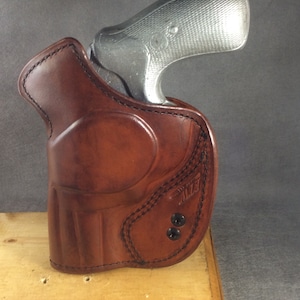 May include: A brown leather holster with a stitched design and two black snaps. A silver revolver is partially visible inside the holster.