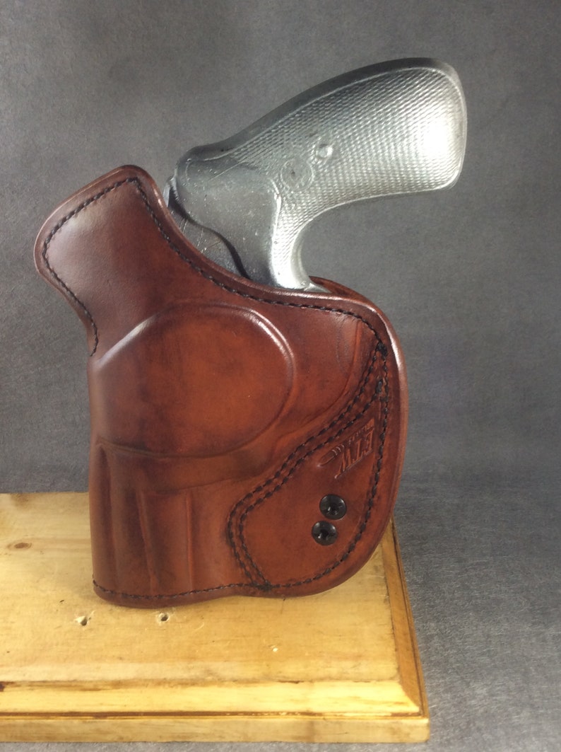 May include: A brown leather holster with a stitched design and two black snaps. A silver revolver is partially visible inside the holster.