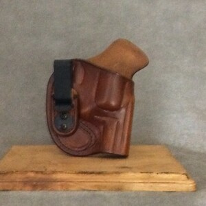 May include: A brown leather holster with a black clip for a handgun. The holster is on a wooden surface.
