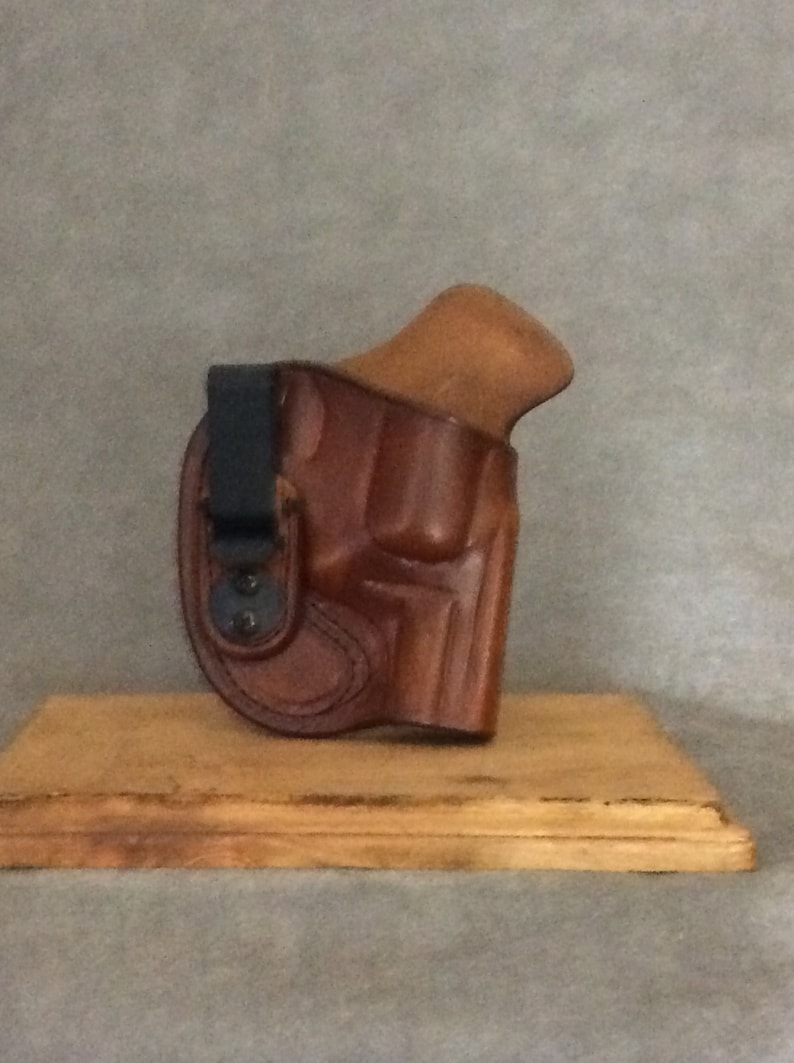 May include: A brown leather holster with a black clip for a handgun. The holster is on a wooden surface.