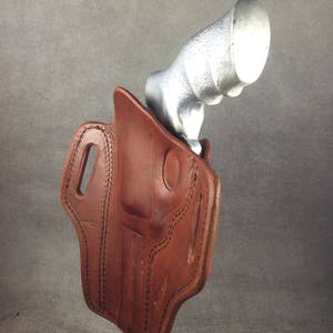 May include: A brown leather holster with stitching detail, designed to hold a silver handgun. The holster is displayed on a stand.