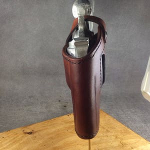 May include: A brown leather holster for a silver handgun. The holster is mounted on a wooden stand.
