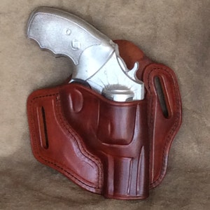 Puede incluir: Una funda de cuero marrón con un revólver plateado en su interior. La funda tiene un cinturón y una correa para asegurar el arma.