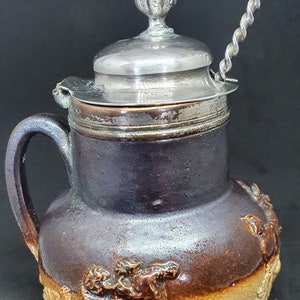 Puede incluir: Una jarra de cerámica marrón con una tapa de plata y un asa de plata. La jarra tiene un diseño decorativo de un caballo y un jinete en el lateral. La jarra mide aproximadamente 20 cm de alto.