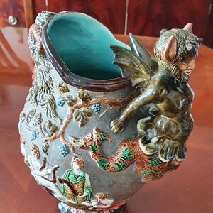 Op de afbeelding: Een decoratieve keramische vaas met een turquoise interieur en een grijze buitenkant. De vaas heeft reliëfsculpturen van figuren, wijnranken en een gevleugelde demon. De basis is donkerbruin.