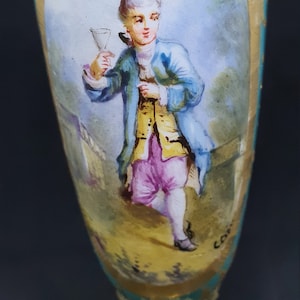 Könnte beinhalten: Dekorative antike Vase mit handgemalter Szene einer Person in Kleidung des 18. Jahrhunderts, die ein Glas hält. Die Vase hat ein türkis-goldenes Farbschema, mit aufwendigen Details und einem Sockel.