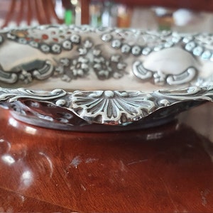 Puede incluir: Un plato de servir ovalado de plata con un diseño ornamentado. El plato tiene un borde elevado con un borde festoneado y intrincados patrones florales.
