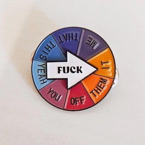 May include: A circular enamel pin featuring a white arrow pointing right with the word FUCK in black text. The background is divided into six colored segments with the words THAT, ME, IT, THEM, OFF, YOU, and YEAH printed in black around the arrow.