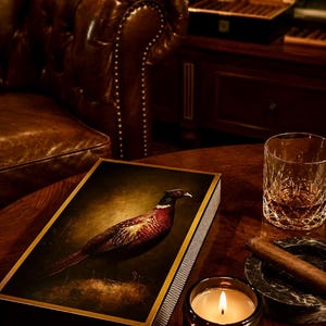 May include: A rectangular box featuring a detailed illustration of a pheasant on its lid, with a gold-toned border and a textured side panel. The box is positioned on a dark wooden surface alongside a glass of amber liquid and a lit candle.