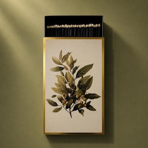 May include: A rectangular matchbox with a gold-trimmed border featuring a botanical illustration of an olive branch with green leaves and small olives on a cream background. The box is partially open, revealing a row of black-tipped wooden matches.