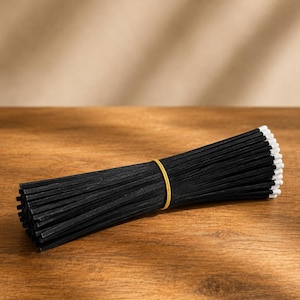 May include: A bundle of long, thin, black wooden matches with white tips, secured in the center by a single yellow rubber band. The matches are arranged horizontally, showcasing their uniform length and contrasting black and white color scheme.