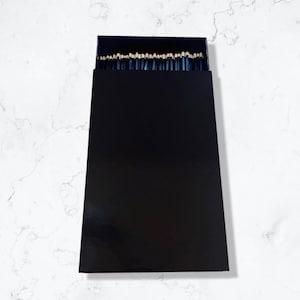 May include: A rectangular black matchbox standing vertically, partially open at the top to reveal a row of dark blue matchsticks with light tan tips arranged in a neat, side-by-side formation against the solid black background of the box.