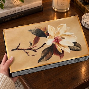 May include: A rectangular decorative box featuring a beige top with a detailed illustration of a white magnolia flower on a brown branch. The box has a gold-toned border and a black base with a textured, silver-colored grid pattern along the side.