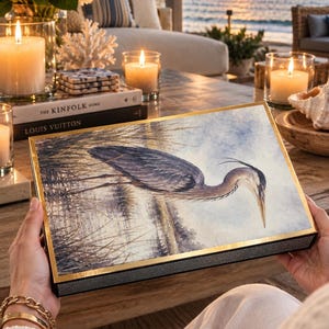 Op de afbeelding: Een rechthoekige decoratieve doos met een illustratie in aquarel-stijl van een grote blauwe reiger in moerasgras. De doos heeft een goudkleurige metalen rand en een zwart paneel met textuur aan de zijkant, met een gedetailleerde vogelscène.