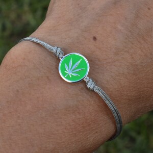 May include: A silver bracelet with a green and white cannabis leaf charm. The charm is round and has a silver border. The bracelet is made of a gray, woven cord. The background is blurred green grass.