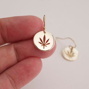 May include: A pair of gold dangle earrings with a circular pendant featuring a cut-out marijuana leaf design.