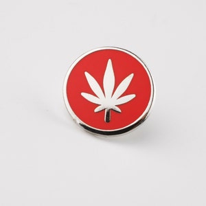 Puede incluir: Pin redondo con borde plateado y fondo rojo. Presenta una hoja de cannabis blanca en el centro. Un peque&ntilde;o accesorio, probablemente para ropa o bolsos.