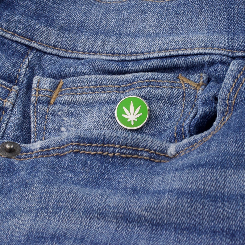 Puede incluir: Un pin redondo de esmalte verde y blanco con un dise&ntilde;o de hoja de cannabis. El pin est&aacute; colocado sobre un fondo de mezclilla azul, descansando en un bolsillo de jeans. El pin tiene un borde blanco y un interior verde.