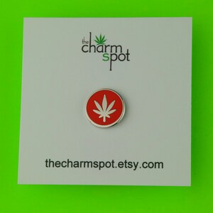 Puede incluir: Un pin redondo, rojo y plateado con un dise&ntilde;o de hoja de cannabis blanca. El pin est&aacute; sobre una tarjeta blanca con el texto "the charm spot" y la URL de la tienda Etsy. El fondo es verde brillante.