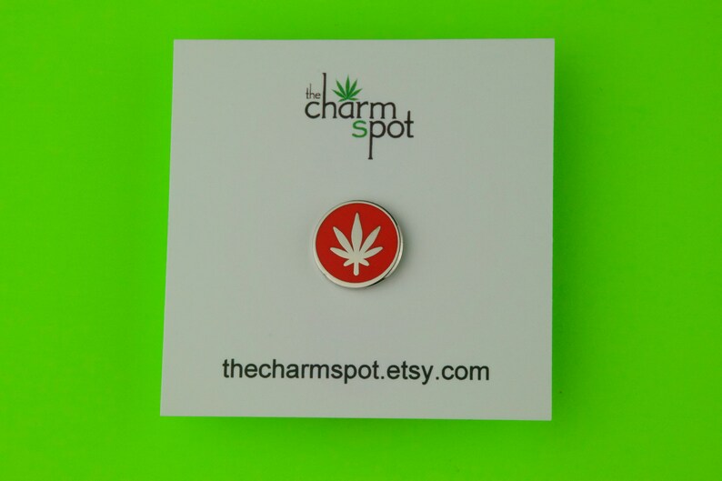 Puede incluir: Un pin redondo, rojo y plateado con un dise&ntilde;o de hoja de cannabis blanca. El pin est&aacute; sobre una tarjeta blanca con el texto "the charm spot" y la URL de la tienda Etsy. El fondo es verde brillante.