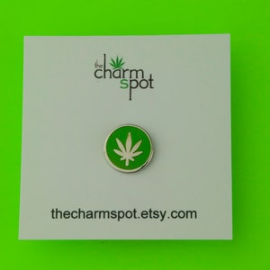 Puede incluir: Un pin redondo, verde y plateado con un dise&ntilde;o de hoja de cannabis blanca. El pin est&aacute; sobre una tarjeta blanca con el texto "the charm spot" y la direcci&oacute;n del sitio web thecharmspot.etsy.com.