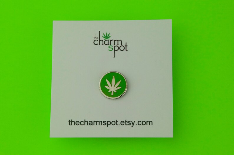 Puede incluir: Un pin redondo, verde y plateado con un dise&ntilde;o de hoja de cannabis blanca. El pin est&aacute; sobre una tarjeta blanca con el texto "the charm spot" y la direcci&oacute;n del sitio web thecharmspot.etsy.com.