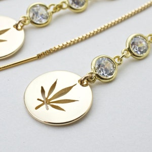 May include: Gold dangle earrings with a round pendant featuring a cut-out marijuana leaf design. The earrings are adorned with clear crystal stones.