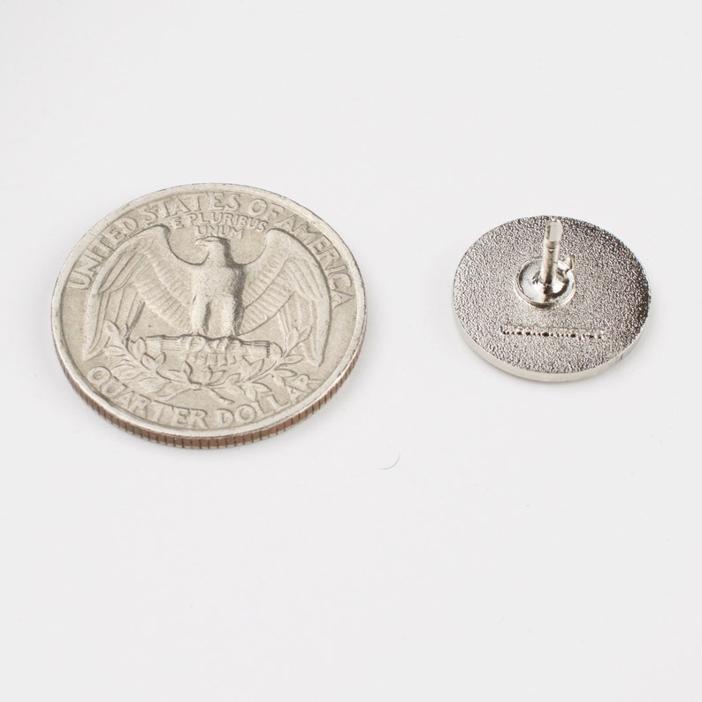 Puede incluir: Un pin redondo plateado con un poste y cierre, junto a una moneda de veinticinco centavos estadounidense. El pin tiene una superficie texturizada y un peque&ntilde;o dise&ntilde;o en relieve. La moneda muestra el perfil de George Washington.