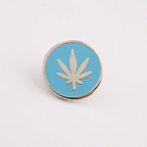 Puede incluir: Un pin redondo plateado con un centro azul claro. El centro presenta un dise&ntilde;o de hoja de cannabis plateada. El pin est&aacute; sobre un fondo blanco.