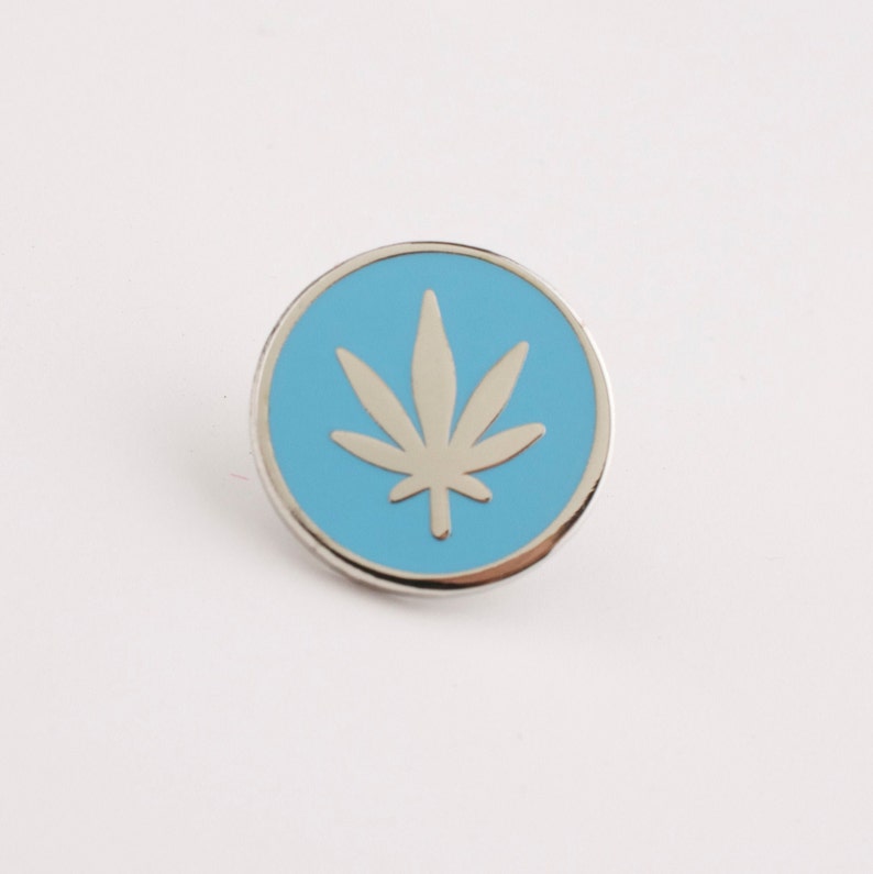 Puede incluir: Un pin redondo plateado con un centro azul claro. El centro presenta un dise&ntilde;o de hoja de cannabis plateada. El pin est&aacute; sobre un fondo blanco.