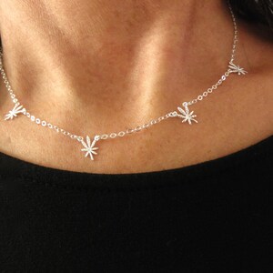 May include: A delicate silver necklace featuring five cannabis leaf charms. The necklace is worn against a black top, highlighting the bright silver of the chain and charms. The charms are evenly spaced along the chain.