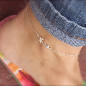 May include: A silver ankle bracelet with a palm tree charm and a small clear stone. The bracelet is on a person's ankle.
