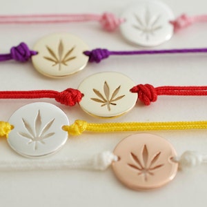 May include: Close-up of several bracelets with a cannabis leaf design. The bracelets feature a circular charm with a cut-out leaf design. The cords are in various colors, including pink, purple, red, yellow, and white. The charms are in gold, silver, and rose gold.