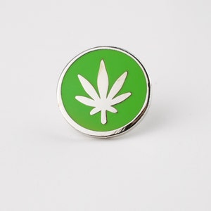 Puede incluir: Un pin redondo plateado con un centro verde brillante. El centro presenta un dise&ntilde;o de hoja de cannabis blanca. El pin est&aacute; sobre un fondo blanco.