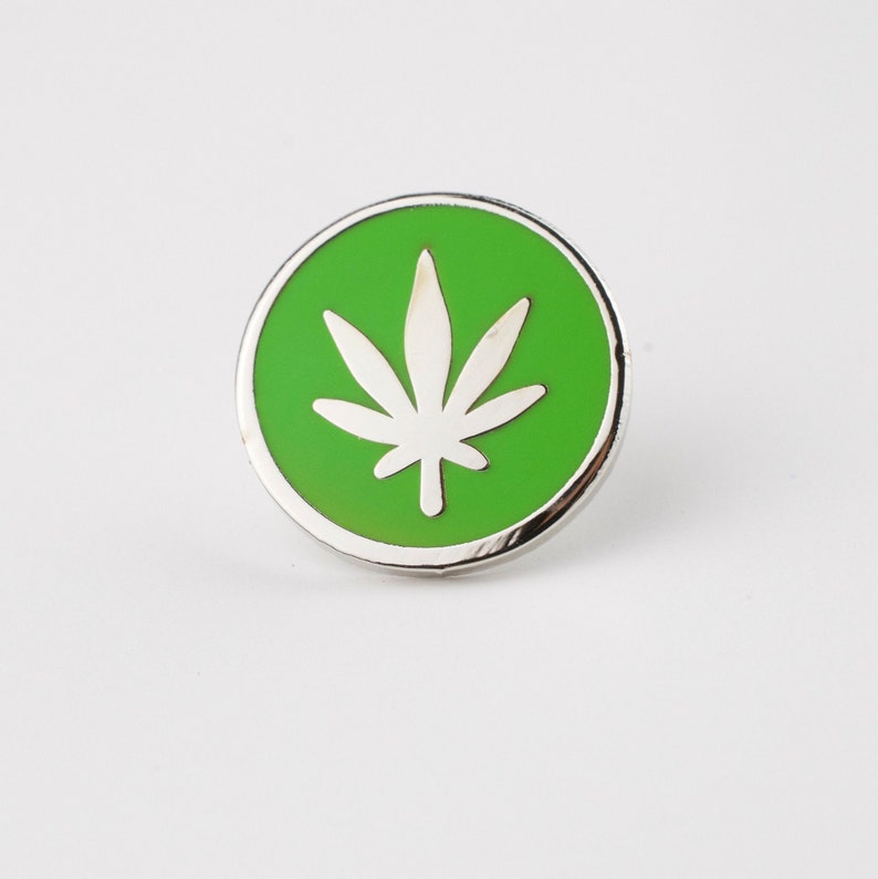 Puede incluir: Un pin redondo plateado con un centro verde brillante. El centro presenta un dise&ntilde;o de hoja de cannabis blanca. El pin est&aacute; sobre un fondo blanco.