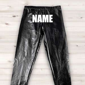 May include: Black leggings with a shiny, faux-leather appearance. The word "NAME" is printed in large, white capital letters across the upper portion of the leggings. The leggings are displayed on a light wood-grain surface.