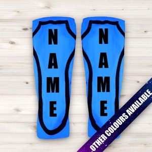 May include: A pair of blue shin guards with black trim and the word "NAME" printed vertically in black on each guard. The text "OTHER COLORS AVAILABLE" is printed on a purple and blue background.