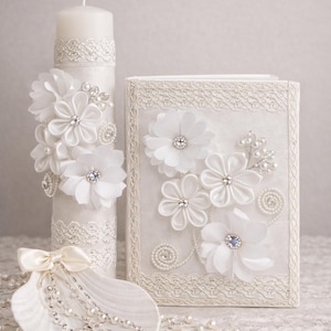 May include: A white candle, book, and seashell with decorative embellishments. The candle and book are adorned with white fabric flowers, pearls, and rhinestones. A seashell holds a rosary with a cross. The items are arranged on a lace surface.