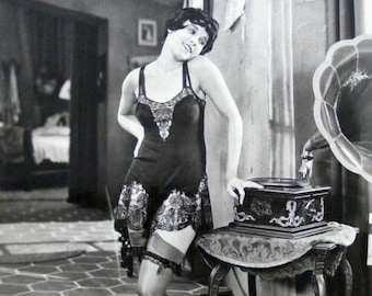 Op de afbeelding: Een vrouw in een zwarte kanten jurk en kousen staat naast een vintage grammofoon. Ze houdt een plaat in haar hand en kijkt naar de grammofoon.