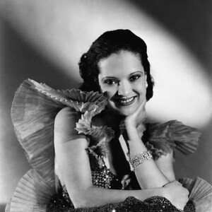 May include: A black and white portrait of a woman wearing a sequined dress with a ruffled neckline. She is smiling and has her hand on her chin.