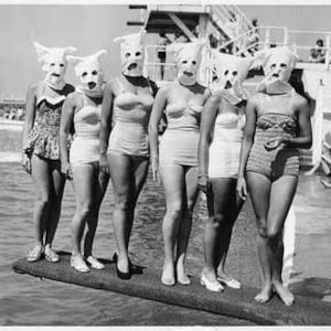 May include: A vintage black and white photograph of six people wearing swimsuits and white masks with eye and mouth openings. They are standing on a diving board near a pool.