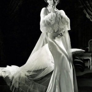 May include: A floor-length white satin gown featuring a fitted silhouette and a sheer, flowing tulle cape with ruffled edges. The garment includes long, voluminous sheer sleeves and a high neckline, captured in a high-contrast black and white photograph.