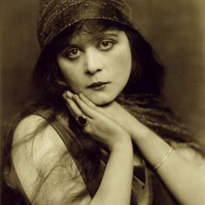 Pode incluir: Retrato em tons sépia de uma mulher com cabelo escuro e um adorno de cabeça com contas. Ela apoia o queixo nas mãos unidas, usando um anel com uma pedra escura e uma pulseira delicada. A imagem tem uma estética vintage, clássica de Hollywood.
